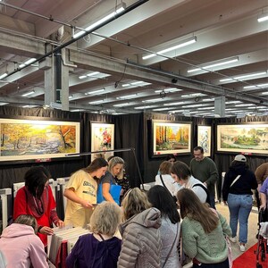 May include: A bustling art fair scene with numerous people examining framed landscape paintings. The paintings feature autumnal scenes and serene landscapes. The fair is held indoors with overhead lighting and a red carpet.