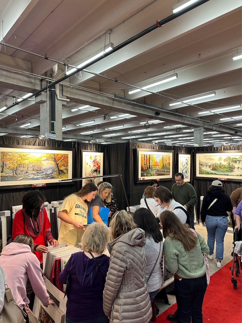May include: A bustling art fair scene with numerous people examining framed landscape paintings. The paintings feature autumnal scenes and serene landscapes. The fair is held indoors with overhead lighting and a red carpet.