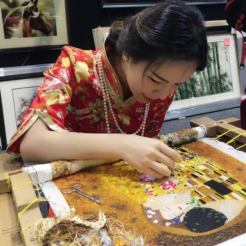 May include: A woman in a red and floral patterned dress is working on a large embroidery project. The embroidery is a recreation of Gustav Klimt's "The Kiss" painting. The woman is using a needle and thread to create the intricate details of the artwork.