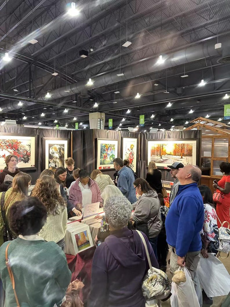 May include: A group of people gathered around a table, looking at framed artwork. The artwork features colorful landscapes and floral designs. The people are browsing through a selection of smaller framed prints.