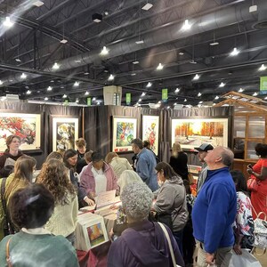 May include: A crowded art fair with numerous people browsing framed artwork. The paintings feature various landscapes and floral themes. The fair is held indoors under bright lighting.