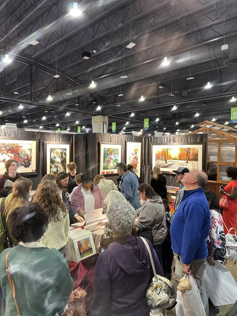 May include: A crowded art fair with numerous people browsing framed artwork. The paintings feature various landscapes and floral themes. The fair is held indoors under bright lighting.