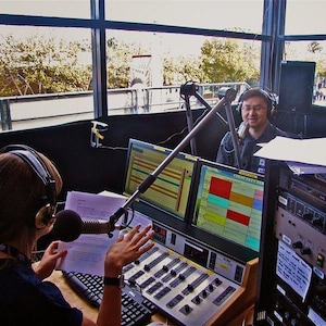 May include: Two people in a radio booth with microphones and headphones. One person is speaking into a microphone, while the other is operating a soundboard. Computer monitors display colorful audio waveforms. The booth has a large window overlooking an outdoor scene.