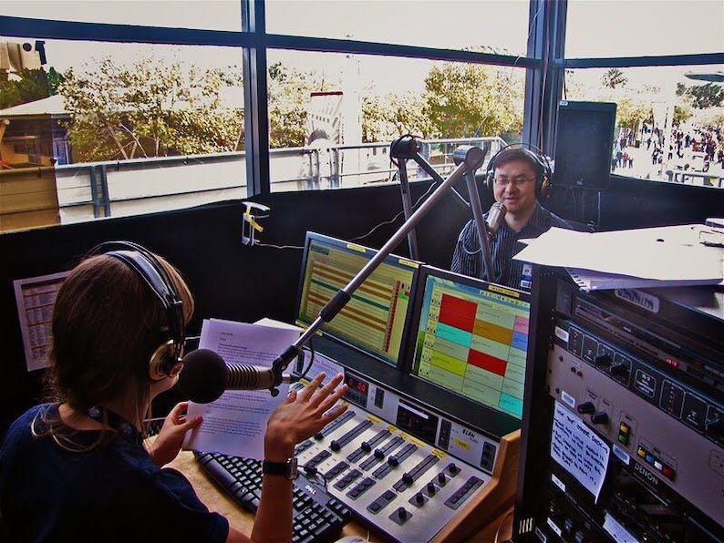 May include: Two people in a radio booth with microphones and headphones. One person is speaking into a microphone, while the other is operating a soundboard. Computer monitors display colorful audio waveforms. The booth has a large window overlooking an outdoor scene.