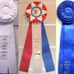 May include: Three award ribbons. The first is white with text "L.A. County Fair" and "Fair's Finest Exhibits 2007". The second is red, white, and blue with "San Diego County Fair" and "Special Award 2007". The third is blue with "First Place" and "2011".