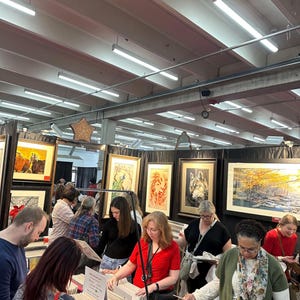 May include: A group of people browse framed artwork at an art fair. The artwork features nature scenes, including a wolf, a bird, and a forest. The artwork is displayed on a wall with white lighting.