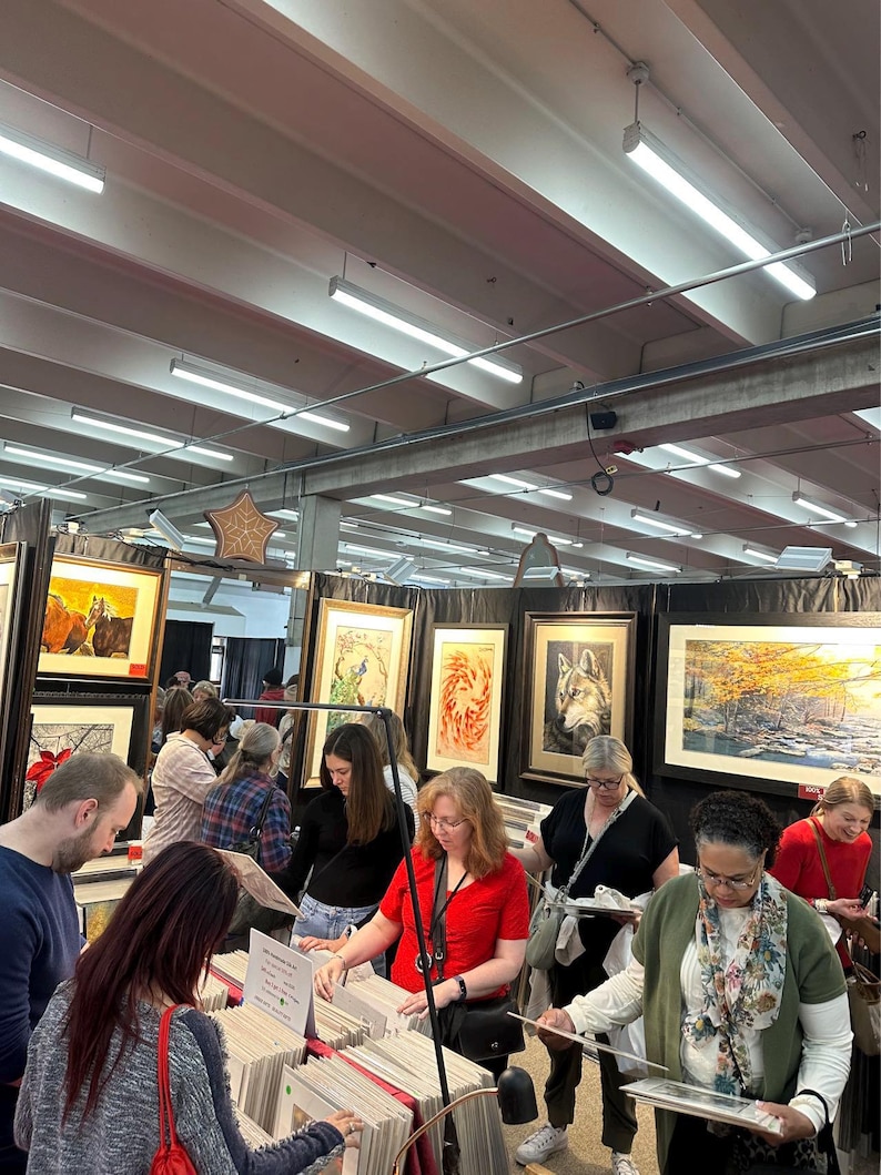 May include: A group of people browse framed artwork at an art fair. The artwork features nature scenes, including a wolf, a bird, and a forest. The artwork is displayed on a wall with white lighting.