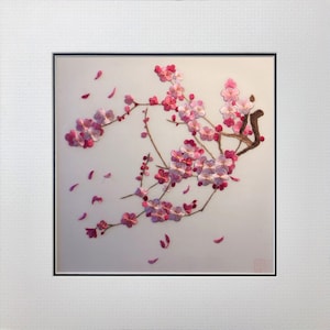 May include: Embroidered artwork of a cherry blossom branch with pink and white flowers and falling petals. The artwork is framed in a white mat and black frame, creating a delicate and elegant aesthetic.