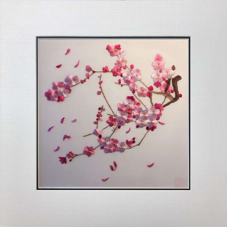 May include: Embroidered artwork of a cherry blossom branch with pink and white flowers and falling petals. The artwork is framed in a white mat and black frame, creating a delicate and elegant aesthetic.