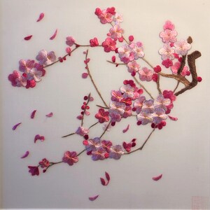 May include: Embroidered artwork featuring a branch of pink and white cherry blossoms with falling petals. The blossoms are detailed with varying shades of pink and white thread, set against a light background. The branches are brown.