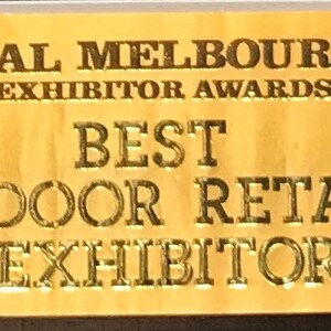 May include: A gold ribbon with the text "2017 ROYAL MELBOURNE SHOW EXHIBITOR AWARDS BEST INDOOR RETAIL EXHIBITOR". The ribbon has a gold fringe and the Royal Melbourne Show logo.