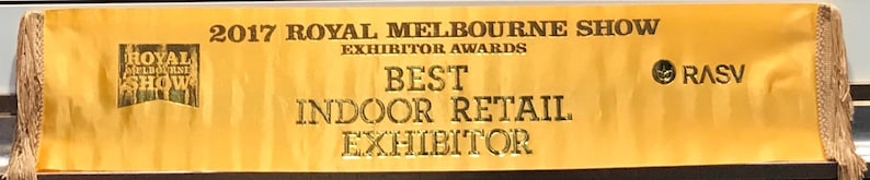 May include: A gold ribbon with the text "2017 ROYAL MELBOURNE SHOW EXHIBITOR AWARDS BEST INDOOR RETAIL EXHIBITOR". The ribbon has a gold fringe and the Royal Melbourne Show logo.