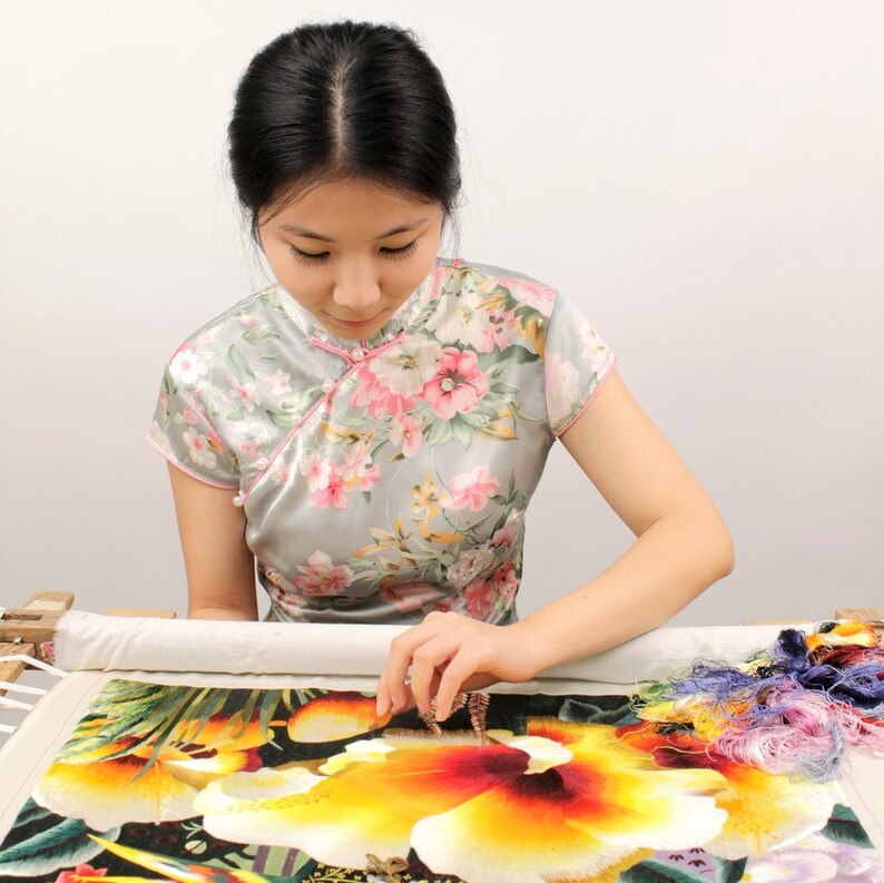 May include: A person working on a colorful floral embroidery project. The person is wearing a gray dress with pink floral patterns. The embroidery features yellow, orange, and red flowers with green leaves. Colorful threads are visible.