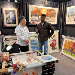 May include: An art fair booth featuring framed artwork and embroidery. Two people are present, one holding a microphone. The artwork includes landscape and floral scenes. Embroidery work is displayed on a wooden frame, with colorful threads and a small lamp.
