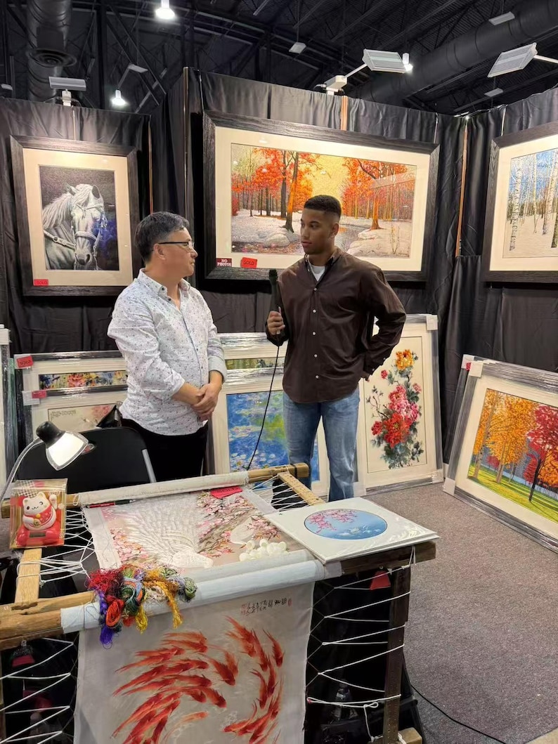 May include: An art fair booth featuring framed artwork and embroidery. Two people are present, one holding a microphone. The artwork includes landscape and floral scenes. Embroidery work is displayed on a wooden frame, with colorful threads and a small lamp.