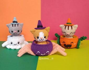 Puede incluir: Tres figuras de gato de crochet para Halloween. El primer gato lleva un sombrero de bruja y está sentado en un recipiente blanco con forma de fantasma. El segundo gato lleva un sombrero de bruja y está sentado en un recipiente morado con forma de caldero. El tercer gato lleva un sombrero de bruja y está sentado en un recipiente naranja con forma de calabaza.