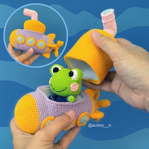 May include: Crocheted toy submarine in yellow, purple, and orange, with a frog passenger. The submarine has portholes and fins. A separate yellow and orange crocheted cylinder is held above the submarine. The frog has rosy cheeks and is wearing a blue shirt.