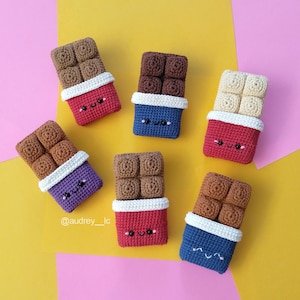 Può includere: Sei barrette di cioccolato all'uncinetto con faccine sorridenti. Ogni barretta presenta quadrati testurizzati marroni o color crema, una fascia bianca e un involucro colorato in rosso, blu o viola. Il testo @audrey__lc è visibile.