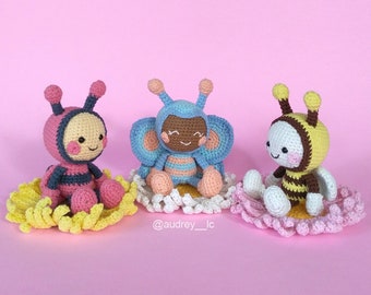 May include: Three crocheted stuffed toys, each sitting on a crocheted flower. The toys are a pink and black bee, a blue butterfly with a brown face, and a yellow and black bee.