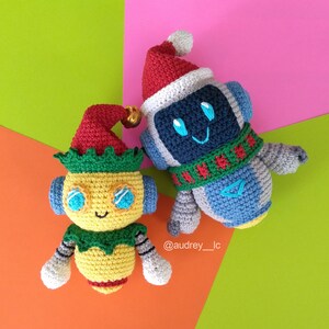 May include: Two crocheted plush toys, one is a robot wearing a red and white Santa hat and a green and red scarf, the other is a yellow elf with a red hat and green trim.  The toys are on a multi-colored background.  @audrey_lc