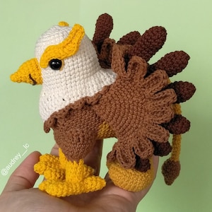 Pode incluir: Um brinquedo de amigurumi de águia crochetado com penas marrons e amarelas. A águia tem uma cabeça branca e um olho preto.