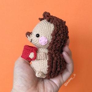 Puede incluir: Un erizo de crochet sosteniendo una taza roja. El erizo es marrón y beige con la nariz rosa.