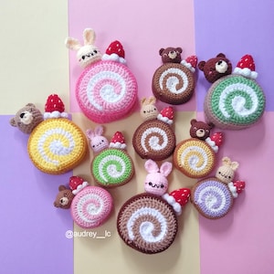 Puede incluir: Una colección de coloridos bizcochos suizos de ganchillo con varios rellenos, cubiertos con pequeños animales de ganchillo y fresas. Los pasteles son de color rosa, marrón, amarillo, verde y morado, con espirales blancas y crema.