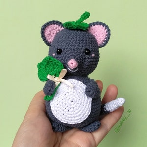 May include: A gray crocheted mouse holding a green shamrock. The mouse has pink ears and a white belly. The mouse is wearing a green crocheted hat.