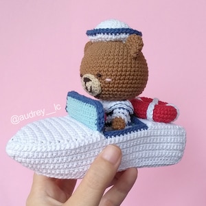 Puede incluir: Un oso de crochet marrón con un gorro de marinero azul y blanco y una camisa a rayas azul y blanco está sentado en un barco de crochet blanco con un asiento a rayas azul y blanco y un salvavidas rojo.