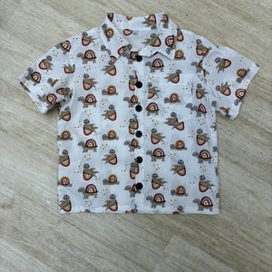 May include: White button-down shirt patterned with turtles and rainbows in brown, red, and yellow. The shirt features short sleeves, a chest pocket, and black buttons. A casual, patterned top.