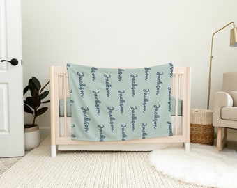 May include: A light green baby blanket with the name "Jackson" repeated in a dark blue script. The blanket is draped over a white crib. The room has a neutral color scheme with a gold lamp and a woven basket.