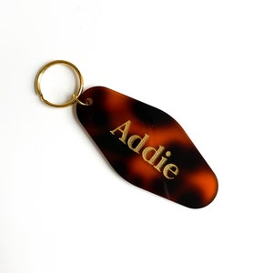 May include: A tortoiseshell-patterned keychain with a gold-colored key ring. The keychain has the name "Addie" in gold lettering. The keychain is a retro motel style.
