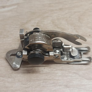 Puede incluir: Un accesorio para máquina de coser Singer, hecho de metal plateado. El accesorio tiene un diseño complejo con varias palancas, tornillos y un botón negro. El logotipo de Singer es visible en el cuerpo principal.