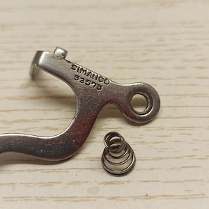 May include: A close-up of two metal sewing machine components. One is a curved lever with the text "SIMANCO 33573" and a circular hole. The other is a small, coiled spring. Both parts are silver and rest on a light wood surface.