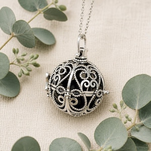May include: A silver-toned spherical locket pendant suspended from a delicate matching chain. The locket features an intricate, openwork filigree design with swirling patterns and beaded textures, revealing a smooth, dark, solid sphere inside.