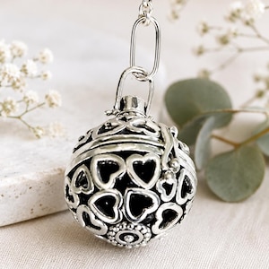 May include: A spherical silver pendant featuring an intricate openwork design of multiple heart shapes. The metal has a darkened finish within the recessed areas to highlight the pattern. It hangs from a silver chain link bail at the top.