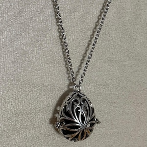 May include: Silver-toned pendant necklace featuring a teardrop-shaped locket with intricate floral filigree. The locket hangs from a delicate chain. The interior of the locket appears dark, contrasting with the bright metal.