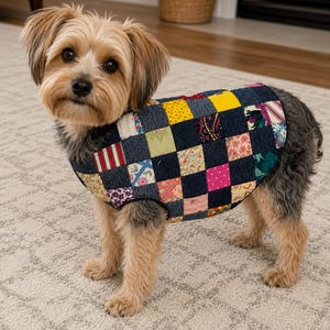May include: A small dog wearing a quilted vest made from a patchwork of dark blue denim squares and various colorful floral, polka dot, and striped fabric squares. The vest features a sleeveless design with black trim around the edges.