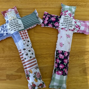 May include: Two handmade fabric crosses with patchwork designs. One cross features blue floral, red gingham, and striped patterns. The other has floral, striped, and gingham patterns. Each cross has a fabric label with text from Mark 11:24.