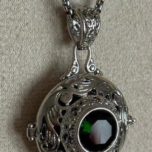 May include: Silver-toned pendant necklace featuring an ornate, filigree-style locket with a faceted, emerald-green gemstone. The locket hangs from a delicate chain. The design includes intricate scrollwork and a secure clasp, creating an elegant accessory.