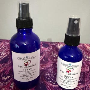May include: Two cobalt blue glass spray bottles with black caps and white labels. The labels read "Pet Calming Spray" with ingredients listed. The bottles are 2 oz and 4 oz. The bottles are on a purple and white patterned surface.