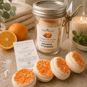 May include: A clear glass mason jar with a silver lid containing round, off-white shower steamers topped with orange crystals. The label reads Sweet Orange Essential Oil SHOWER STEAMERS. Includes a white instruction card and four loose steamers.