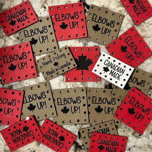 May include: A collection of red, tan, and white square tags with black text and a maple leaf design. The tags read "ELBOWS UP!" and "CANADIAN MADE." Some tags have small holes around the edges.
