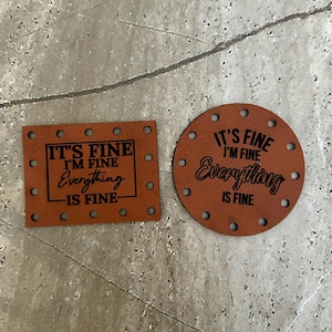 Puede incluir: Dos parches de cuero marrón con texto negro. El primer parche es rectangular y dice "It's Fine I'm Fine Everything Is Fine". El segundo parche es redondo y dice "It's Fine I'm Fine Everything Is Fine".