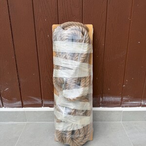 May include: A rectangular wooden board wrapped in clear plastic film, featuring a thick, coiled natural fibre rope secured to the surface with multiple horizontal strips of white adhesive tape, creating a compact and bundled appearance.