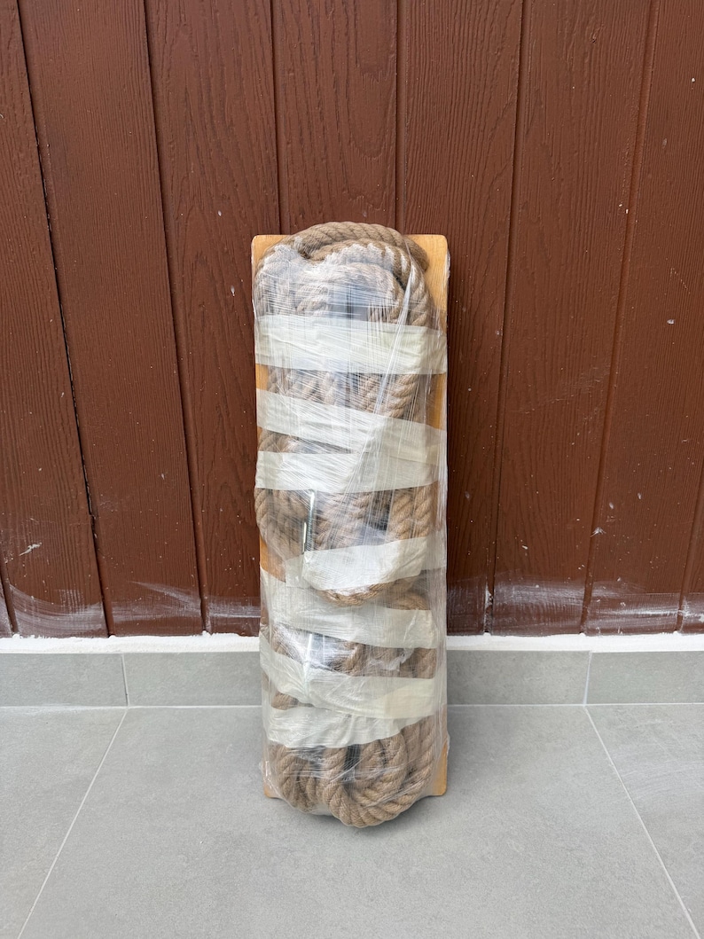 May include: A rectangular wooden board wrapped in clear plastic film, featuring a thick, coiled natural fibre rope secured to the surface with multiple horizontal strips of white adhesive tape, creating a compact and bundled appearance.