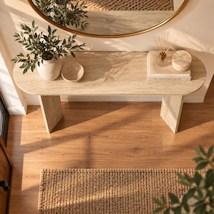 May include: A beige travertine console table with a smooth oval top and two sturdy slab legs. The table surface holds a ceramic vase with green foliage, a small bowl, a glass reed diffuser, and a book titled The Kinfolk Home with a round stone box.