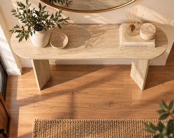 May include: A beige travertine console table with a smooth oval top and two sturdy slab legs. The table surface holds a ceramic vase with green foliage, a small bowl, a glass reed diffuser, and a book titled The Kinfolk Home with a round stone box.