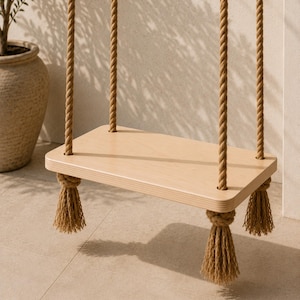 May include: A rectangular light-coloured wooden swing seat suspended by four thick, twisted tan jute ropes. Each rope passes through a corner hole in the seat and is secured underneath with a large, decorative knot that ends in a frayed tassel.