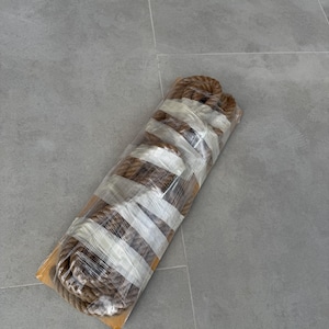 May include: A coiled length of thick, brown, natural fibre rope secured with clear plastic wrap and white adhesive tape. The rope is wound into a cylindrical shape and rests on a flat, rectangular cardboard base, showing a textured surface.